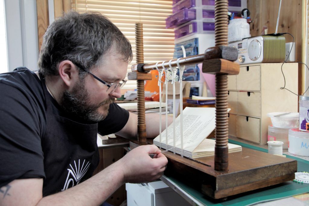 bookbinder sewing a book on four tapes on a wooden sewing frame