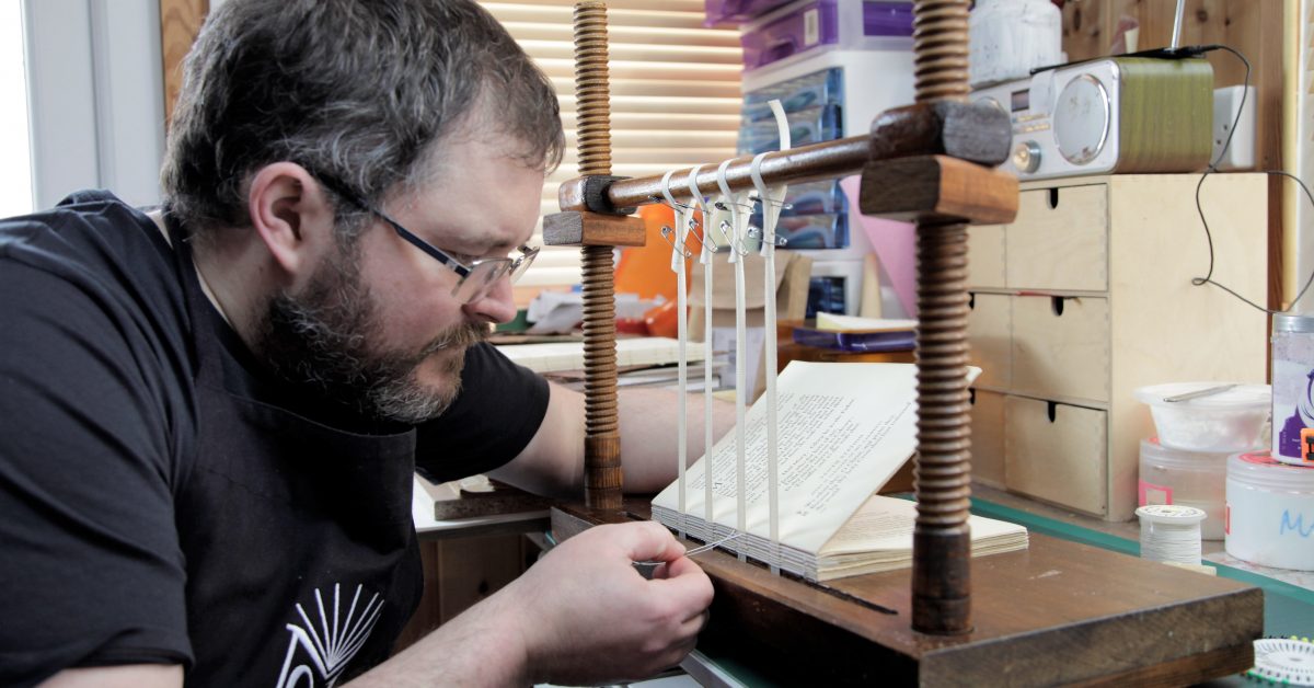 bookbinder sewing a book on four tapes on a wooden sewing frame
