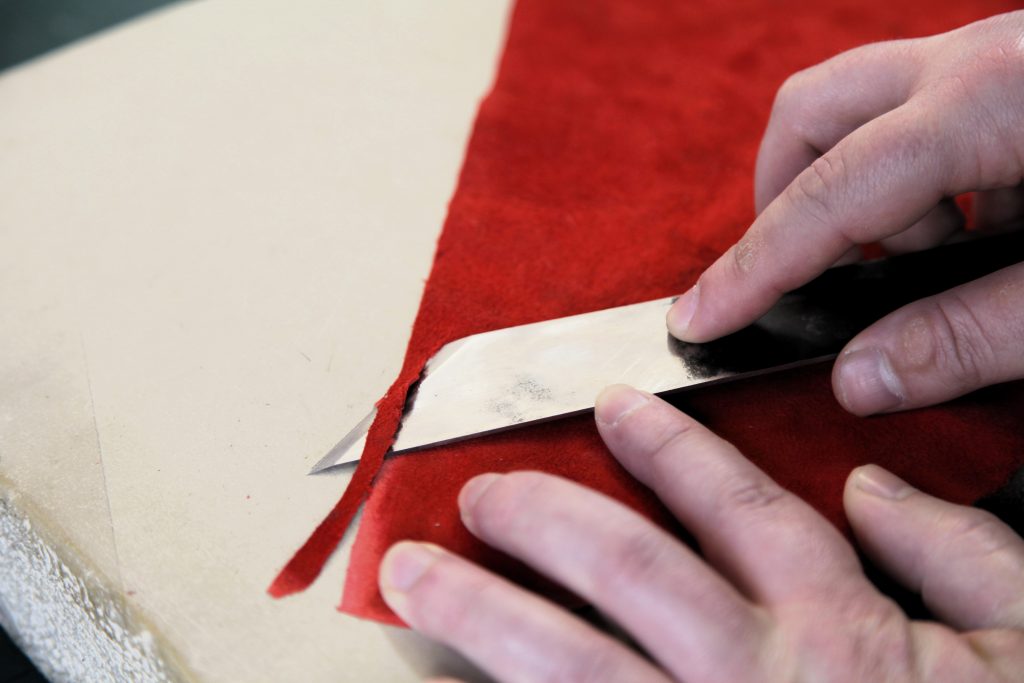 closeup of hands using an English paring knife, edge paring a piece of red leather on a lithostone